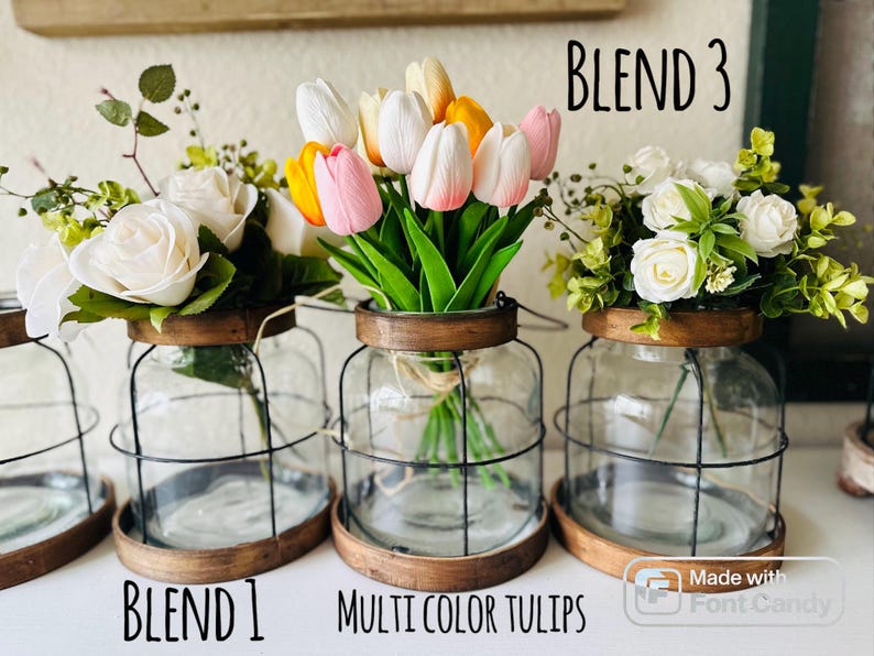 K&ouml;nnte beinhalten: Drei Glasvasen mit Drahtk&auml;figen, jede gef&uuml;llt mit verschiedenen Blumenarrangements. Die erste Vase enth&auml;lt wei&szlig;e Rosen und Gr&uuml;nzeug, die zweite enth&auml;lt mehrfarbige Tulpen und die dritte enth&auml;lt wei&szlig;e Rosen und Gr&uuml;nzeug. Die Vasen sind mit "BLEND 1", "MULTI COLOR TULIPS" und "BLEND 3" beschriftet.