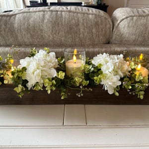 May include: A wooden box centerpiece with white hydrangea flowers, green foliage, and three lit candles in glass holders.