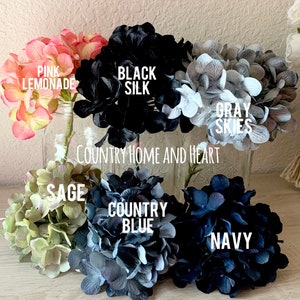 May include: Six bunches of faux hydrangea flowers in various colors, including pink lemonade, black silk, gray skies, sage, country blue, and navy. The flowers are arranged in a row on a wooden surface. The text "COUNTRY HOME AND HEART" is visible below the flowers.