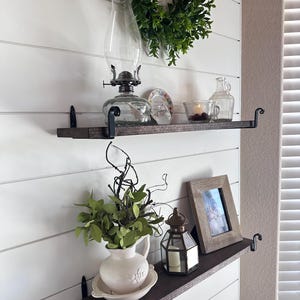 May include: Two dark wooden shelves with black metal brackets are mounted on a white wall. The top shelf holds a glass oil lamp, a small decorative plate, and a glass container. The bottom shelf displays a white pitcher with greenery, a lantern, and a framed photo.