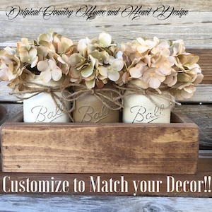 Op de afbeelding: Drie witte geverfde Ball-glazen met beige kunstmatige hortensia bloemen, gerangschikt in een houten doos met een rustieke bruine vlek. De doos heeft de tekst "Customize to Match your Decor!" gedrukt op de voorkant.
