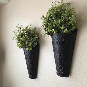 May include: Two black metal wall planters with a decorative floral pattern. Each planter holds a small green bush.