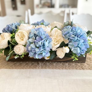 K&ouml;nnte beinhalten: Ein rechteckiger Holzkasten mit k&uuml;nstlichen Blumen. Das Arrangement zeigt hellblaue Hortensien, cremefarbene Pfingstrosen und wei&szlig;e Rosen, akzentuiert mit gr&uuml;nem Laub. Der Mittelpunkt steht auf einem beigen Tischl&auml;ufer.