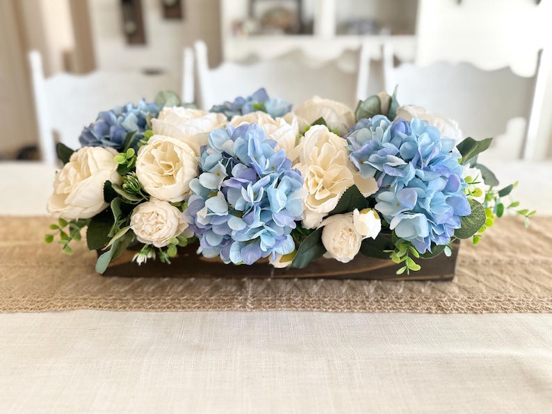 K&ouml;nnte beinhalten: Ein rechteckiger Holzkasten mit k&uuml;nstlichen Blumen. Das Arrangement zeigt hellblaue Hortensien, cremefarbene Pfingstrosen und wei&szlig;e Rosen, akzentuiert mit gr&uuml;nem Laub. Der Mittelpunkt steht auf einem beigen Tischl&auml;ufer.