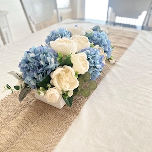 K&ouml;nnte beinhalten: Rechteckiges wei&szlig;es Blumenarrangement mit blauen Hortensien und cremefarbenen Rosen. Die Blumen sind in einem wei&szlig;en rechteckigen Beh&auml;lter angeordnet, der auf einem beigefarbenen Tischl&auml;ufer aus Jute &uuml;ber einer wei&szlig;en Tischdecke platziert ist. Die Szene spielt in einem hellen Raum.
