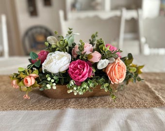 May include: A wooden bowl filled with a floral arrangement of pink, white, and peach roses. The bowl is sitting on a burlap runner on a white table.
