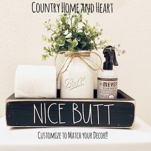 May include: Black wooden box with the text "NICE BUTT" in white lettering. The box contains a roll of toilet paper, a white mason jar with greenery, and a bottle of hand soap. The box is sitting on a white surface. The text "CUSTOMIZE TO MATCH YOUR DECOR!!" is below the box.
