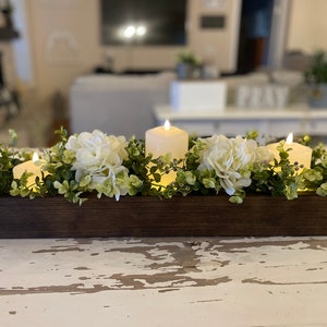 Puede incluir: Un centro de mesa de caja de madera con flores de hortensia blancas y velas encendidas. La caja est&aacute; llena de vegetaci&oacute;n y las velas est&aacute;n rodeadas de flores.