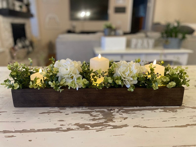 Puede incluir: Un centro de mesa de caja de madera con flores de hortensia blancas y velas encendidas. La caja est&aacute; llena de vegetaci&oacute;n y las velas est&aacute;n rodeadas de flores.