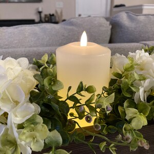 Puede incluir: Una vela blanca en forma de pilar con una llama parpadeante se encuentra en una caja de madera rodeada de hortensias blancas y follaje verde.