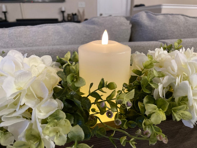 Puede incluir: Una vela blanca en forma de pilar con una llama parpadeante se encuentra en una caja de madera rodeada de hortensias blancas y follaje verde.