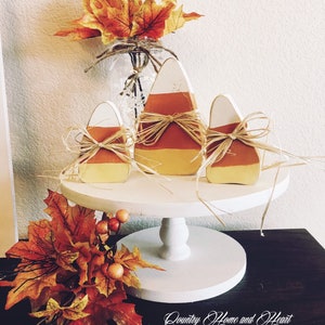 May include: Three wooden candy corn decorations on a white cake stand with fall leaves and berries. The candy corn is painted in orange, yellow, and white. The stand is on a dark wood surface. "Country Home and Heart" is written on the surface.
