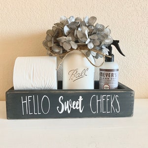 May include: A black wooden box with the text "Hello Sweet Cheeks" on the front. The box contains a roll of toilet paper, a white jar with the word "Ball" written on it, and a bottle of Meyer's Clean Day spray.