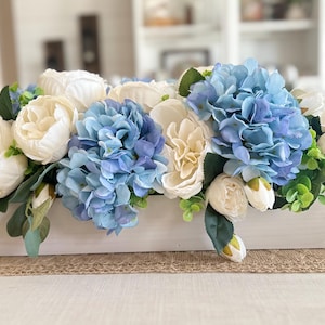K&ouml;nnte beinhalten: Eine rechteckige, wei&szlig;e Holzkiste, &uuml;berf&uuml;llt mit k&uuml;nstlichen Blumen. Das Arrangement zeigt gro&szlig;e blaue Hortensien, cremefarbene Pfingstrosen und wei&szlig;e Bl&uuml;ten, akzentuiert mit gr&uuml;nem Laub. Die Kiste steht auf einem Jutel&auml;ufer.
