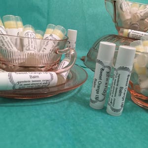 May include: A collection of lip balms in clear tubes, displayed in pink teacups and saucers. The lip balms are labeled "Sweet Orange Lip Balm" and "Lemon Lip Balm". The background is a teal color.