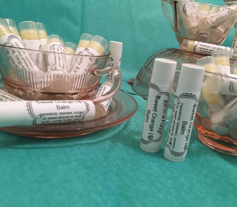 May include: A collection of lip balms in clear tubes, displayed in pink teacups and saucers. The lip balms are labeled "Sweet Orange Lip Balm" and "Lemon Lip Balm". The background is a teal color.