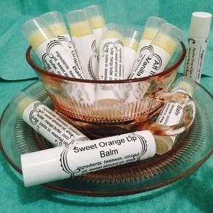 May include: A collection of lip balms in white tubes with labels, displayed in a pink glass teacup and saucer. The labels feature text like "Sweet Orange Lip Balm" and "Vanilla." The balms are arranged on a teal surface.