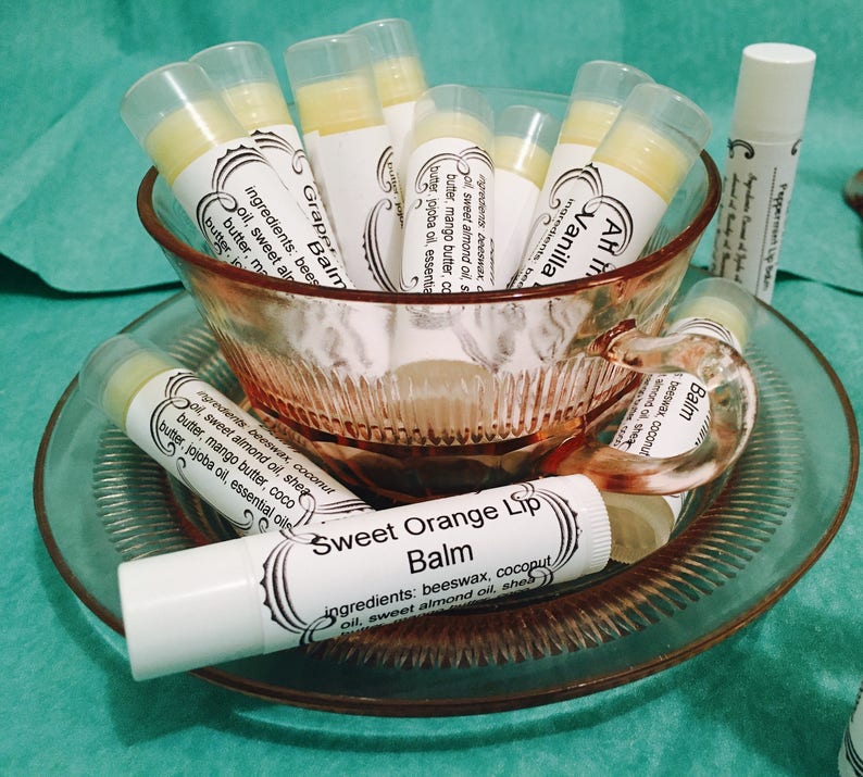 May include: A collection of lip balms in white tubes with labels, displayed in a pink glass teacup and saucer. The labels feature text like "Sweet Orange Lip Balm" and "Vanilla." The balms are arranged on a teal surface.