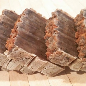 May include: Four bars of handmade soap with a brown and gray marbled appearance. The soap bars are stacked on top of each other and have a rough, textured surface.