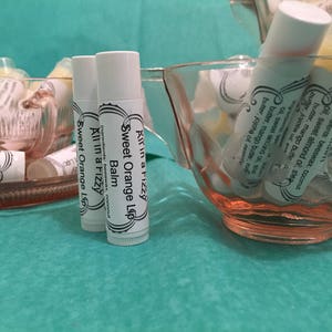 May include: White lip balm tubes with the text "Sweet Orange Lip Balm" are displayed in pink glass bowls. The lip balms are labeled "All in a Fizzy" and list ingredients. The background is a teal-colored surface.
