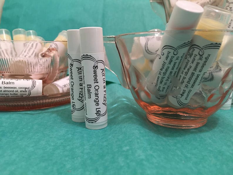 May include: White lip balm tubes with the text "Sweet Orange Lip Balm" are displayed in pink glass bowls. The lip balms are labeled "All in a Fizzy" and list ingredients. The background is a teal-colored surface.
