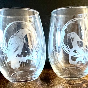 May include: Two clear glass wine glasses with etched designs of ravens in a circle. The ravens are facing opposite directions.