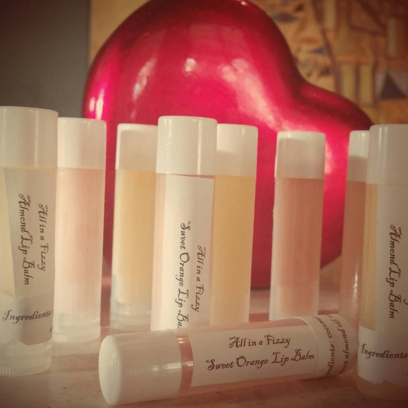 May include: A collection of lip balms with white caps and labels. The labels read "All in a Fizzy" and "Sweet Orange Lip Balm" or "Almond Lip Balm". The lip balms are arranged in front of a large, red heart-shaped object.