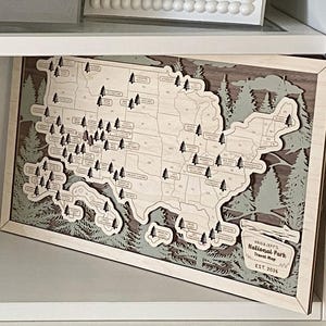 May include: A layered wooden map of the United States featuring cutouts for national parks marked with small tree icons. The map includes a plaque with the text "Kais & Jeff's National Park Travel Map EST. 2026" and is framed in light wood.