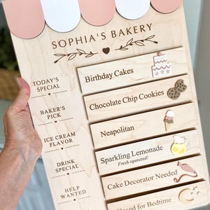 May include: A wooden bakery sign featuring a pink and white scalloped awning, the text "SOPHIA'S BAKERY," and six interchangeable horizontal slats. The slats display "Birthday Cakes," "Chocolate Chip Cookies," "Neapolitan," "Sparkling Lemonade," "Cake Decorator Needed," and "Closed for Bedtime" with corresponding icons.