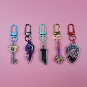 May include: Five acrylic keychains with various designs, including a key, a figure, a sword, a musical instrument, and a shield. Each keychain has a different colored clasp: blue, black, teal, gold, and red. The background is a solid pink.