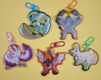 May include: A collection of five acrylic keychains featuring colorful monster designs. Each keychain has a different monster illustration with a unique color scheme and a metal clasp for attachment. The keychains are displayed on a yellow background.