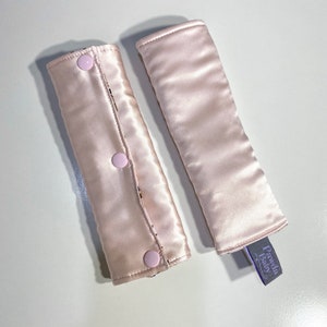 May include: Two light pink satin seatbelt covers. One cover is open, revealing a patterned interior and three pink buttons. The other cover is closed. A small purple tag with the text "Pawda Baby" is attached to one cover.