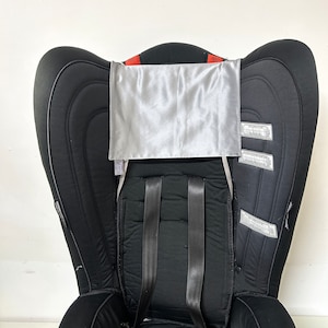 May include: A black car seat with a gray satin headrest cover. The seat has a five-point harness and is designed for a child.