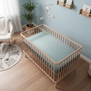 May include: A light wood baby crib with a mint green fitted sheet. The crib has a slatted design and rounded corners. A mobile with clouds and moons hangs above the crib. The room has light blue walls and hardwood floors.