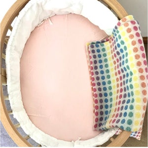 May include: A light wood baby bassinet with a pink fitted sheet, white bumper, and a rainbow polka dot blanket. The bassinet has a round shape.
