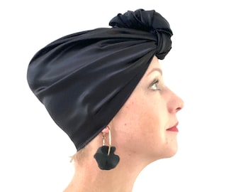 May include: A black satin turban with a knot on top. The turban is worn by a person with black earrings.