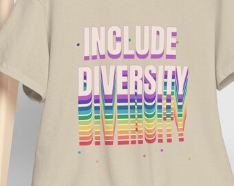 Può includere: Una maglietta beige con la scritta "INCLUDE DIVERSITY" in un design a strati e colorato. La maglietta è appesa a un gancio di legno, con un berretto da baseball bianco appoggiato su una sedia di legno sullo sfondo.