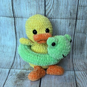 May include: A yellow crocheted duck holding a green crocheted inner tube. The duck has black eyes and an orange beak.