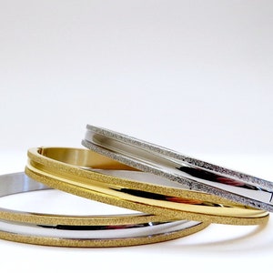 May include: Three stacked bangle bracelets with a textured finish. The top bracelet is silver, the middle bracelet is gold, and the bottom bracelet is silver.