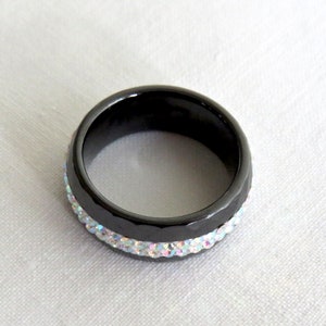 Puede incluir: Un anillo negro con una banda de piedras de imitaci&oacute;n iridiscentes.