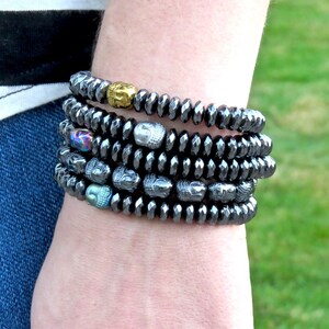 May include: A stack of five black hematite bracelets with silver and gold Buddha head beads. The bracelets are worn on a person's wrist.