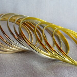 May include: A collection of gold and silver bangles arranged on a gray fabric. The bangles are thin and oval-shaped, reflecting light. The gold bangles have a bright, shiny finish, while the silver ones have a more muted sheen. The jewelry is displayed in a close-up shot.