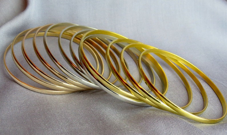 May include: A collection of gold and silver bangles arranged on a gray fabric. The bangles are thin and oval-shaped, reflecting light. The gold bangles have a bright, shiny finish, while the silver ones have a more muted sheen. The jewelry is displayed in a close-up shot.