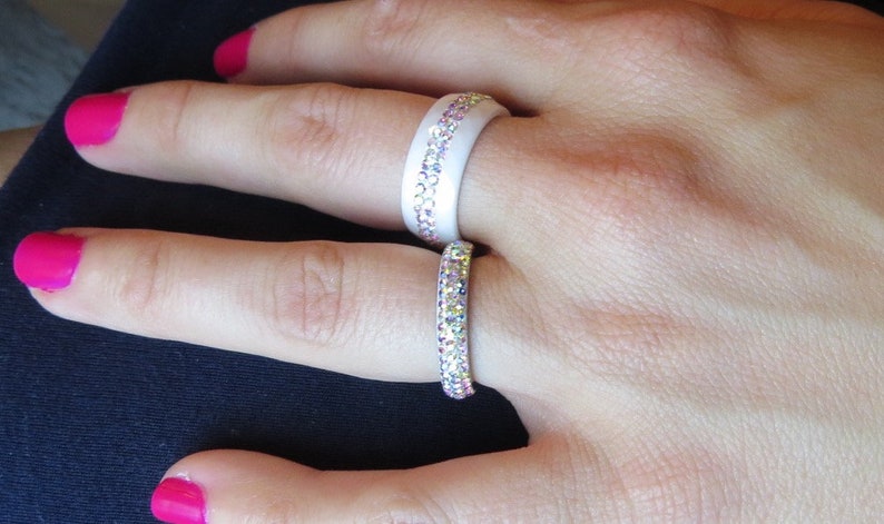Puede incluir: Dos anillos de silicona blancos con filas de piedras de cristal iridiscentes. Los anillos se llevan en el &iacute;ndice y el dedo medio de una mano.