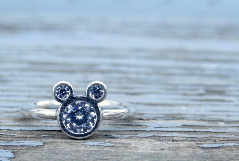 May include: A silver ring with a clear crystal Mickey Mouse head design. The ring has two smaller crystals on the ears.
