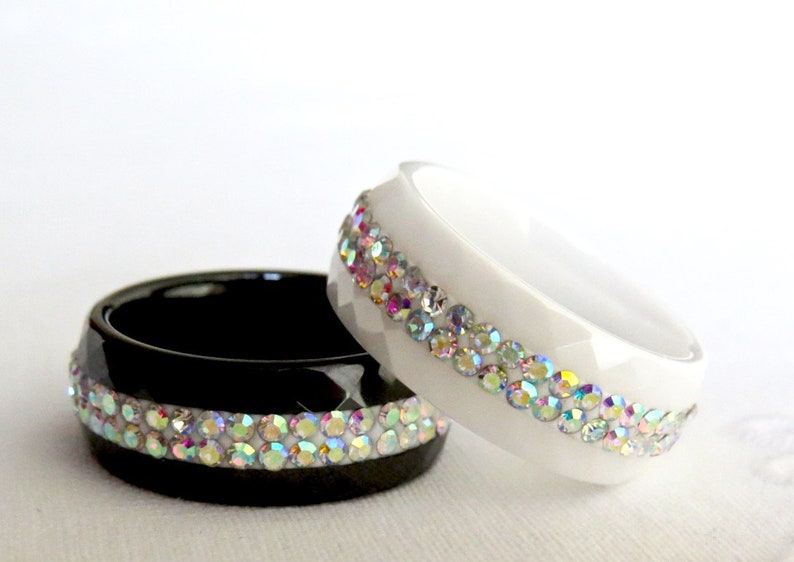 Puede incluir: Dos anillos con una fila de piedras de cristal iridiscentes. Un anillo es negro y el otro es blanco.