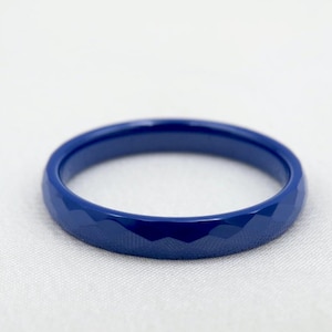 Puede incluir: Un anillo facetado de color azul cobalto. El anillo tiene un diseño geométrico con múltiples superficies planas. El anillo está sobre un fondo blanco.