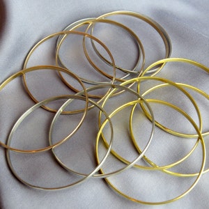 May include: A collection of gold and silver-toned bangles arranged on a gray fabric. The thin, circular bracelets are intertwined, creating a layered effect. The jewelry is displayed against a soft, neutral background.