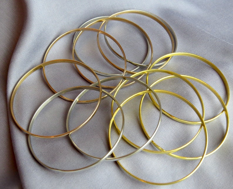 May include: A collection of gold and silver-toned bangles arranged on a gray fabric. The thin, circular bracelets are intertwined, creating a layered effect. The jewelry is displayed against a soft, neutral background.