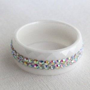 Puede incluir: Anillo de servilleta de cer&aacute;mica blanca con una banda de piedras de imitaci&oacute;n iridiscentes alrededor del borde superior.